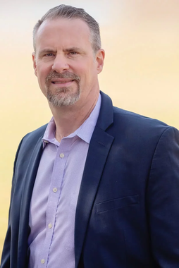 A man in a suit and a purple shirt is standing in front of a yellow background.