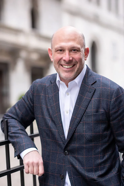 A man in a suit is leaning against a fence and smiling.
