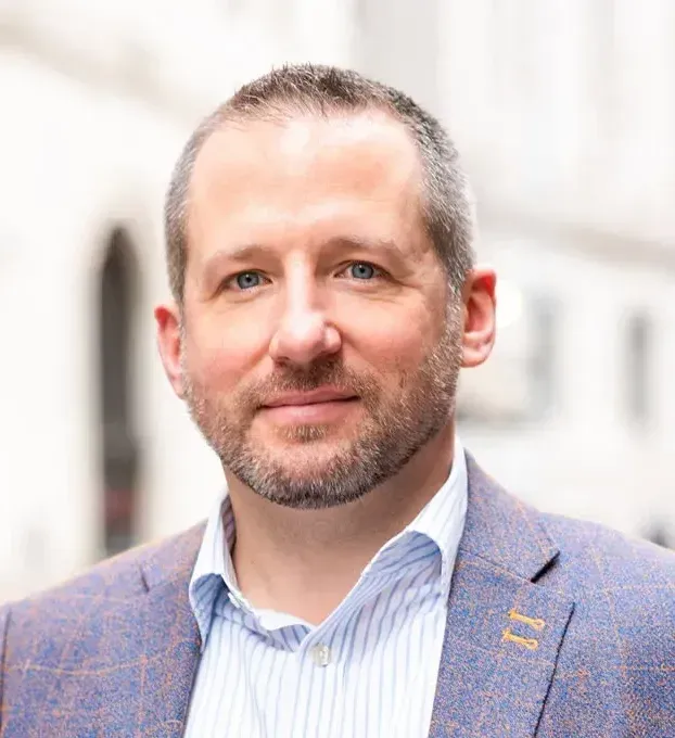 A man in a suit and striped shirt is standing in front of a building.