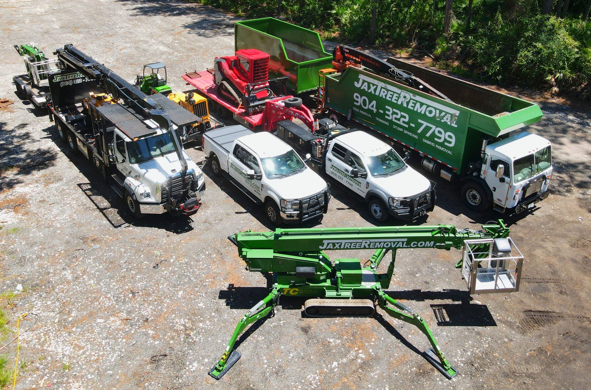 Vehicles of Jax Tree Removal: a green dump truck, two white trucks, and heavy equipment on a trailer under a cloudy sky.