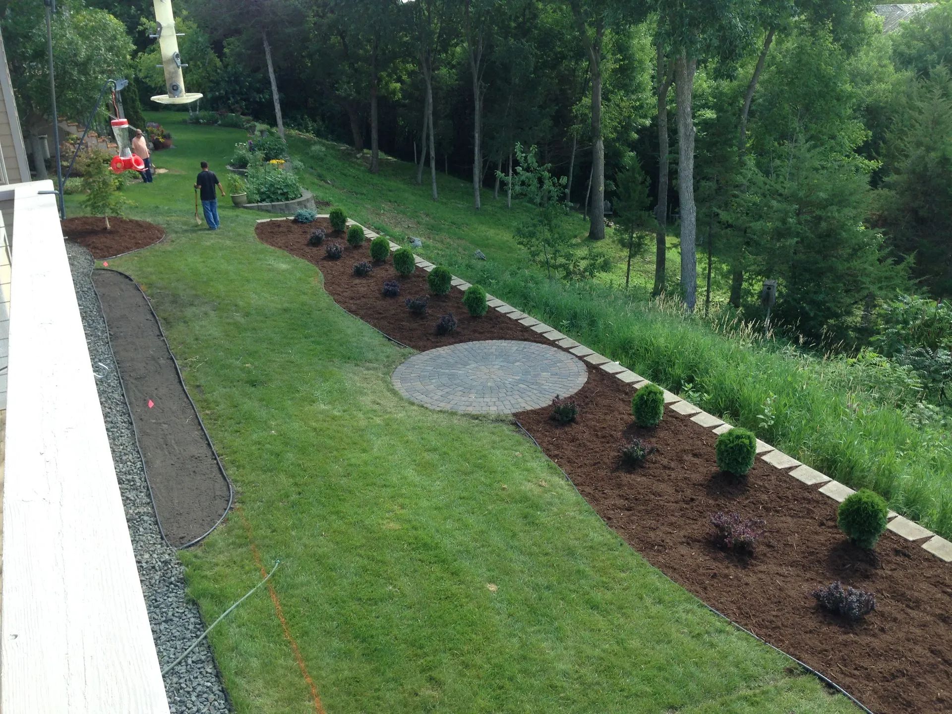 A lawn with a curved mulch bed containing shrubs, a circular patio, a stone border, and a person walking in the distance.