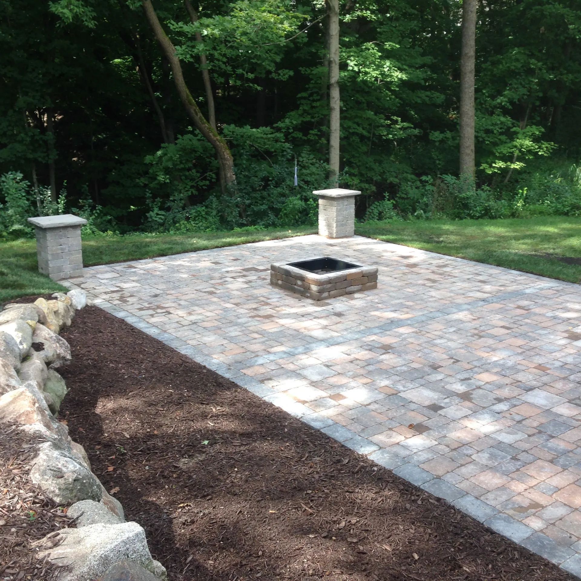 Patio with a fire pit, two pillars, and a mulched border.  Green trees are in the background.