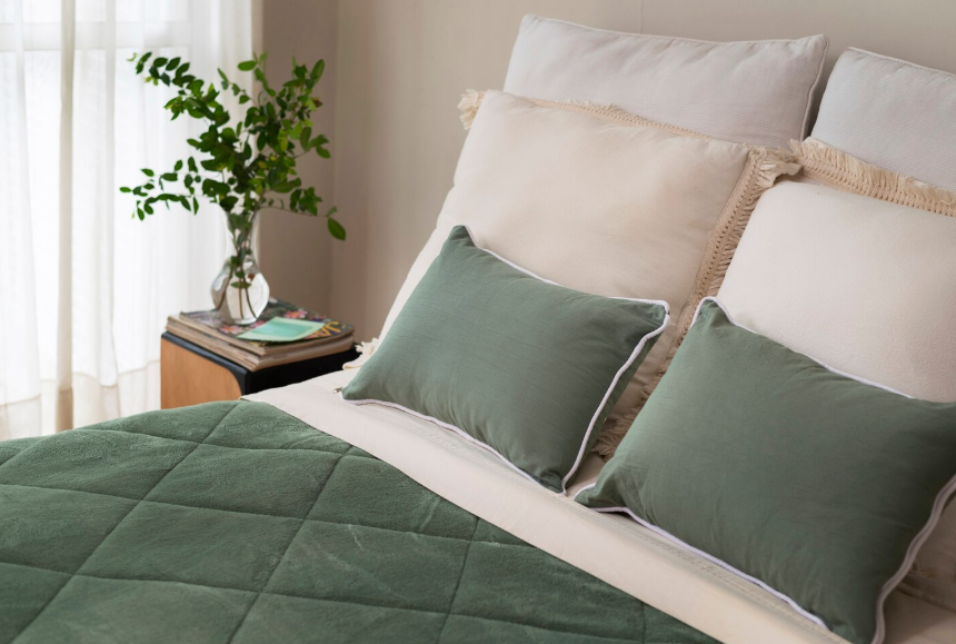 Cama con edredón verde, almohadas y mesita con jarrón y vegetación.