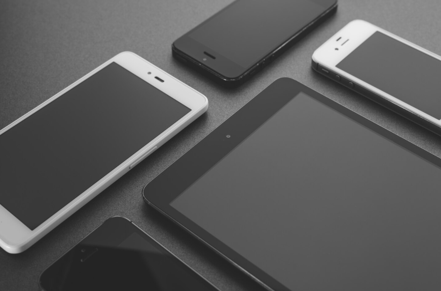 Fotografía en blanco y negro de una tableta y unos teléfonos inteligentes, dispuestos sobre una superficie oscura.