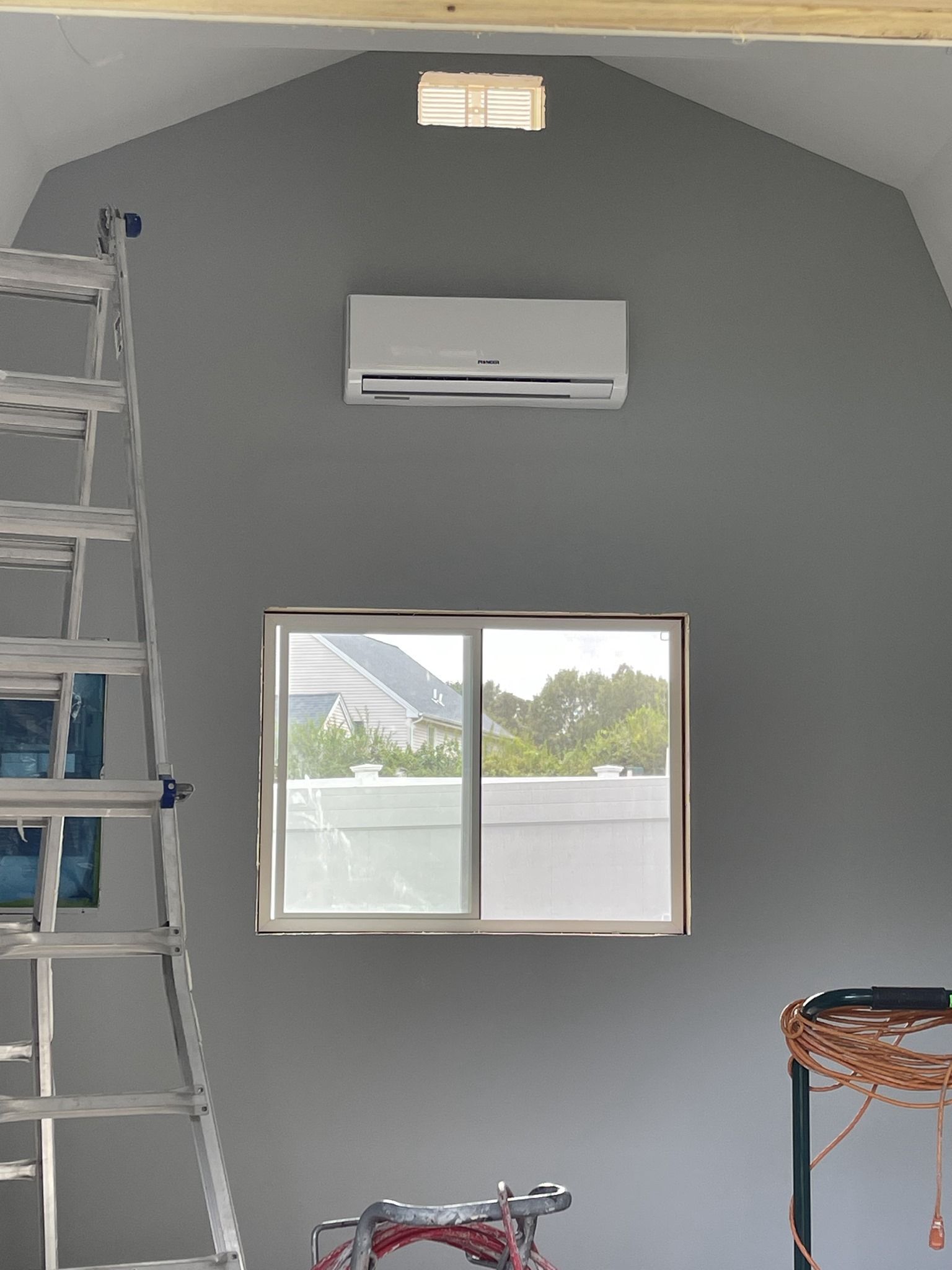 A room with a ladder , a window , AC and a ceiling fan.