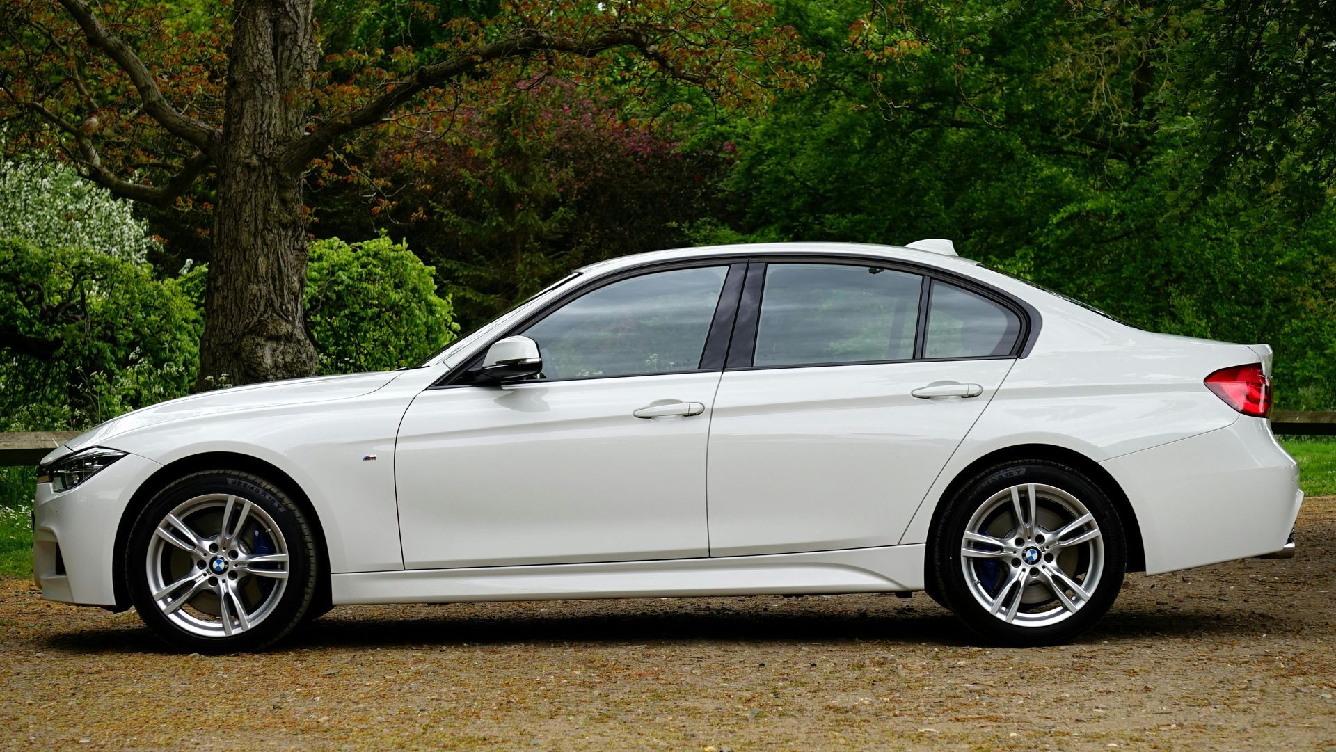 A white car is parked in a gravel lot in front of trees.