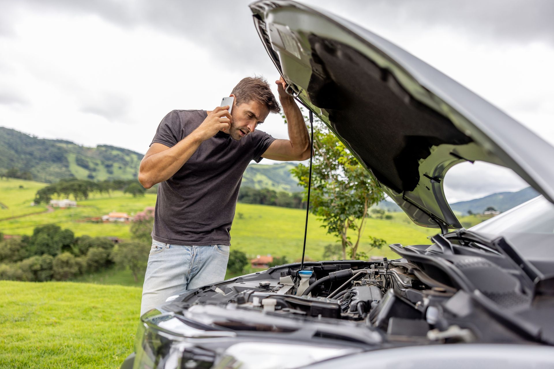 A man is talking on a cell phone while looking under the hood of his broken down car.