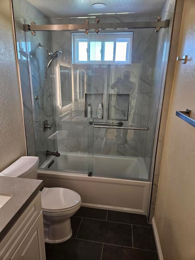 A bathroom featuring a white toilet, a grey vanity, and a walk-in shower with a glass sliding door and marble-style tile.