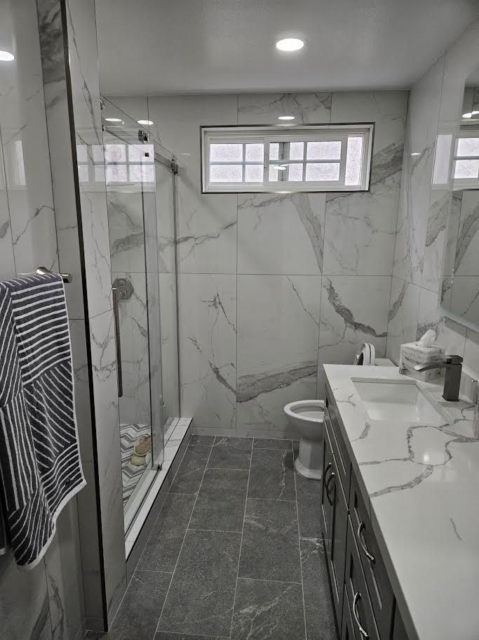 A modern bathroom with white marble-patterned tile walls, a glass walk-in shower, a stone vanity, and a gray tiled floor.