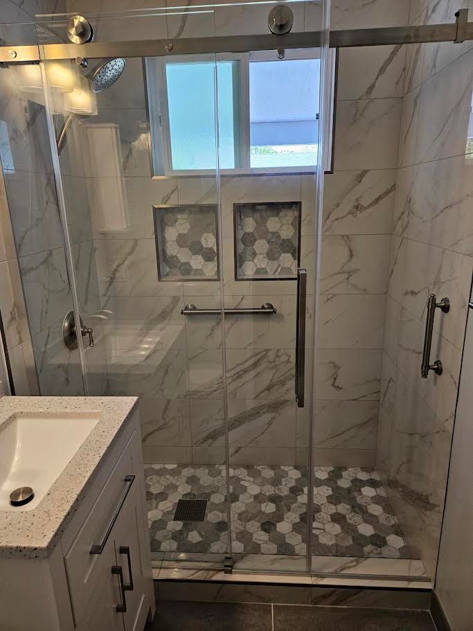 A modern walk-in shower with white marble-patterned tiles, a gray hexagon tile floor, dual wall niches, and a glass door.