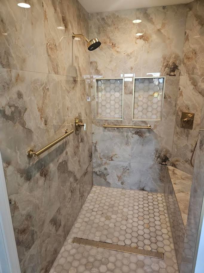 A walk-in shower with marble-look walls, a hexagonal tiled floor, gold fixtures, two recessed niches, and a corner bench.