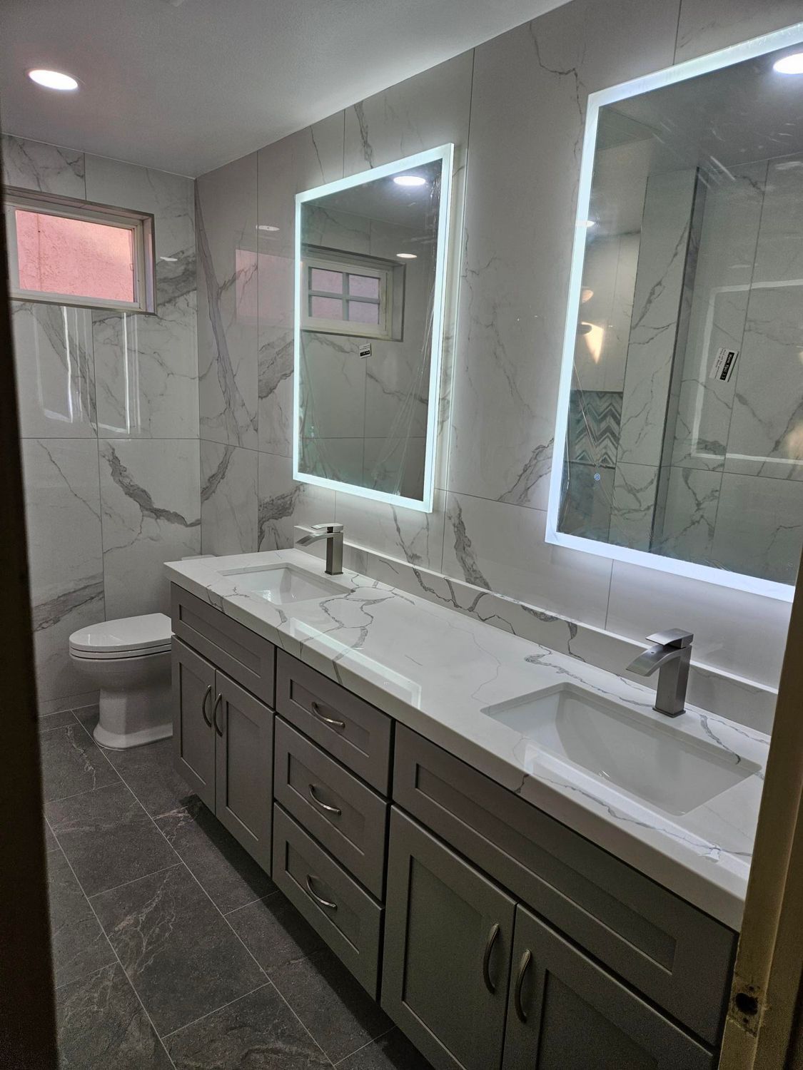 Modern bathroom with a double vanity, gray cabinets, marble-look tiled walls, and two illuminated mirrors.