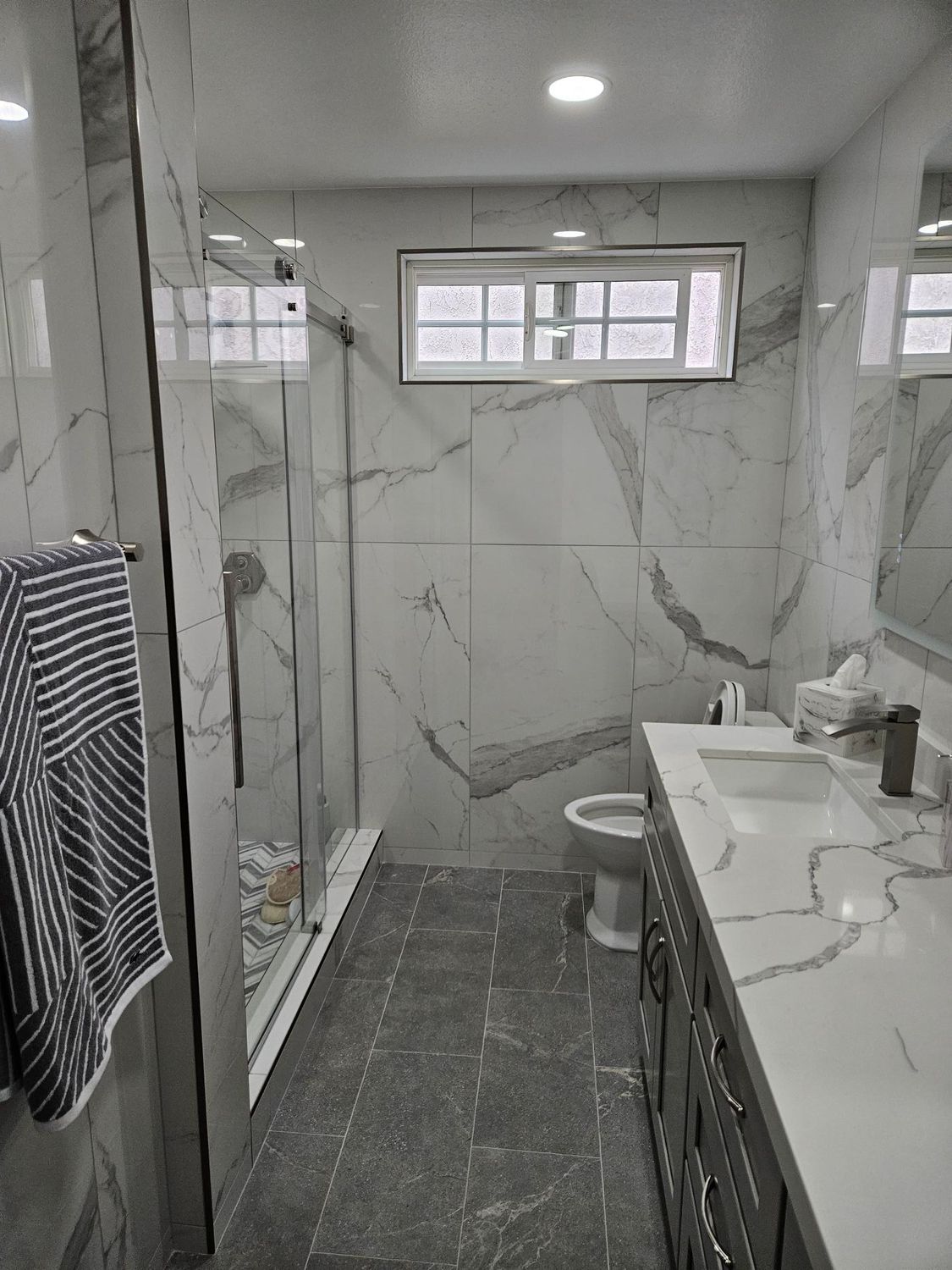 A modern bathroom with a glass-enclosed shower, marble-patterned tile walls, gray tiled floors, and a double vanity.