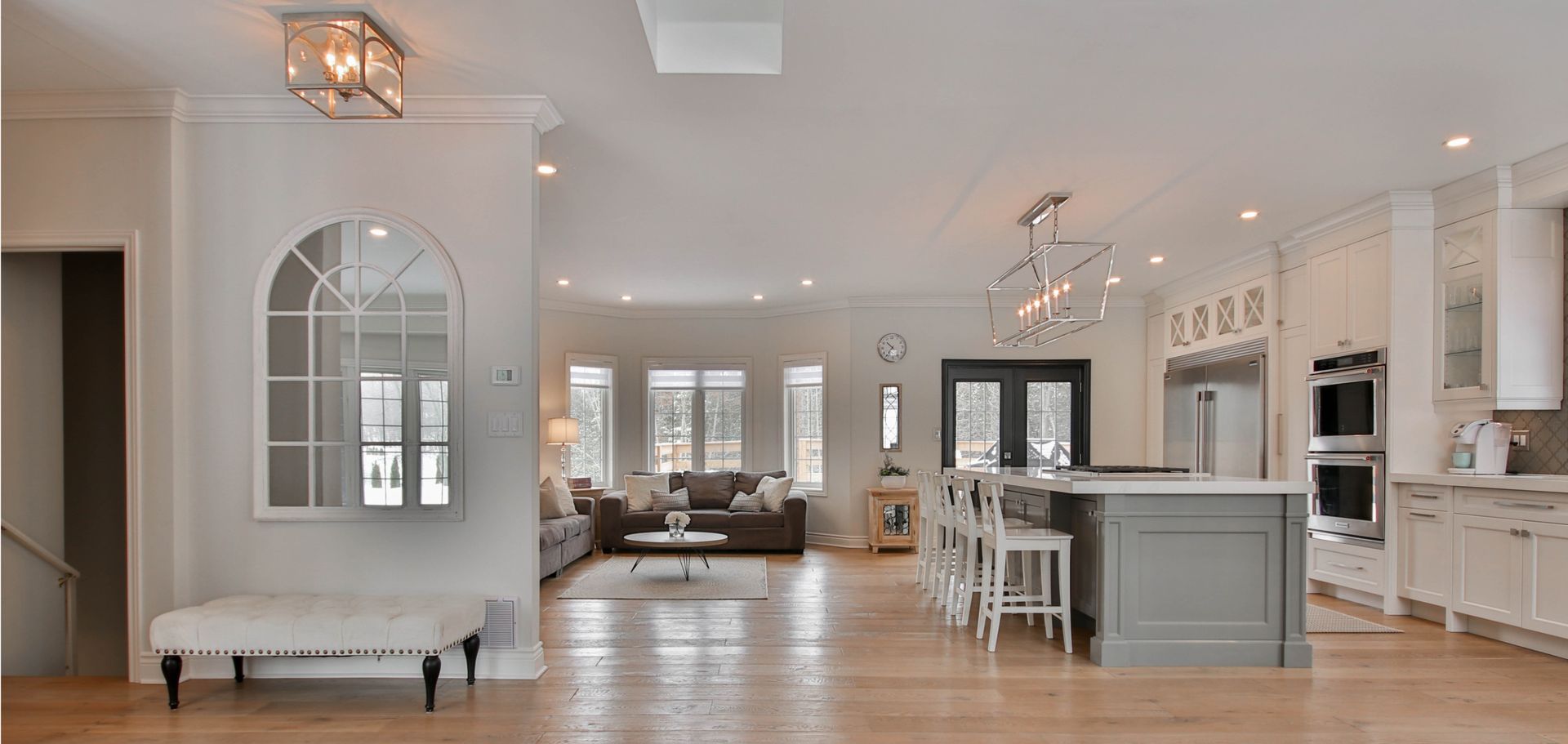 A modern open-plan home featuring a white kitchen island, living room furniture, and arched hallway mirror on wood floors.