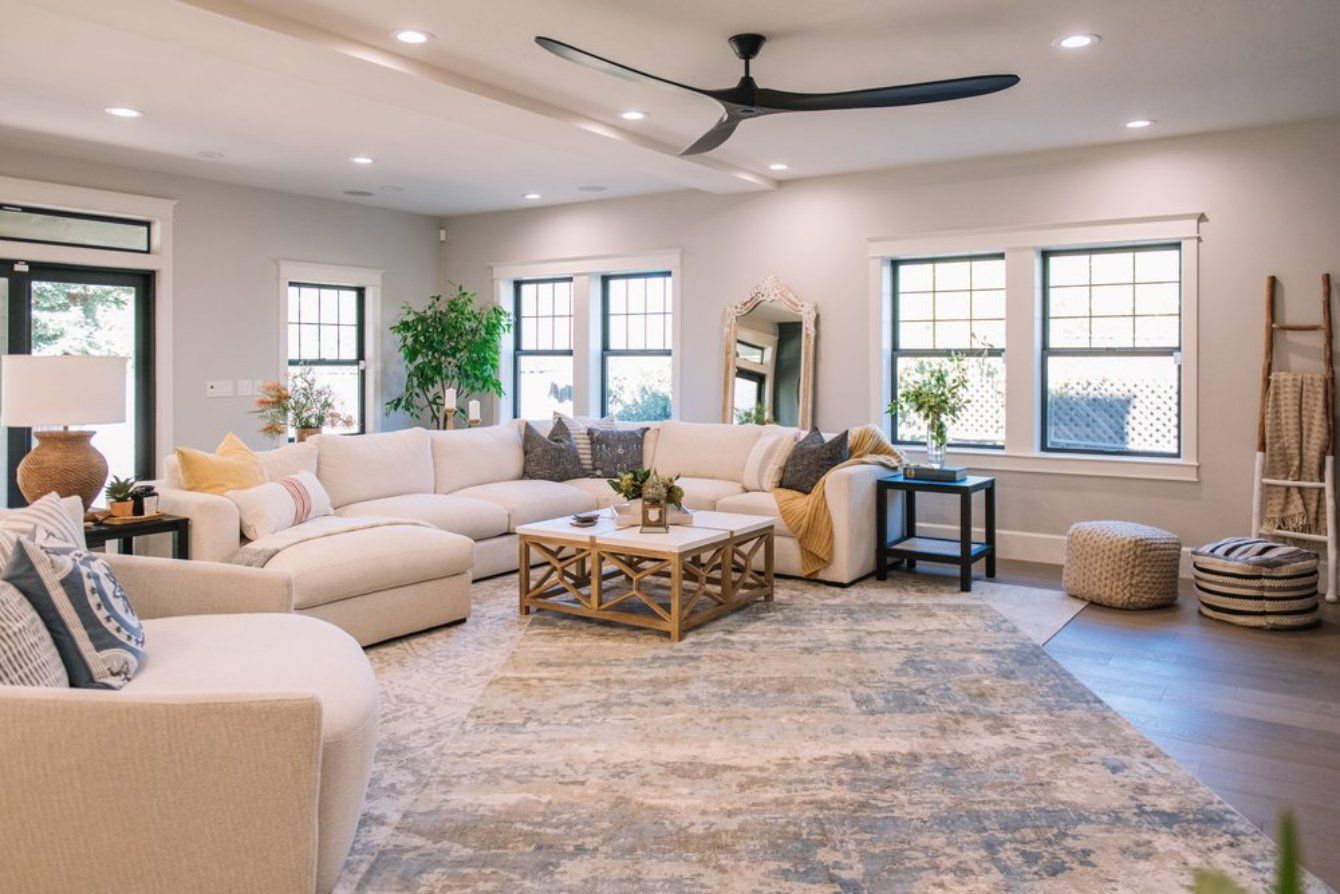 A bright living room features a large cream sectional sofa, an area rug, a coffee table, and a ceiling fan.