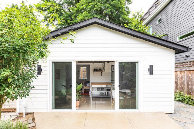 A white, gabled backyard studio with sliding glass doors opening to a concrete patio, framed by greenery.