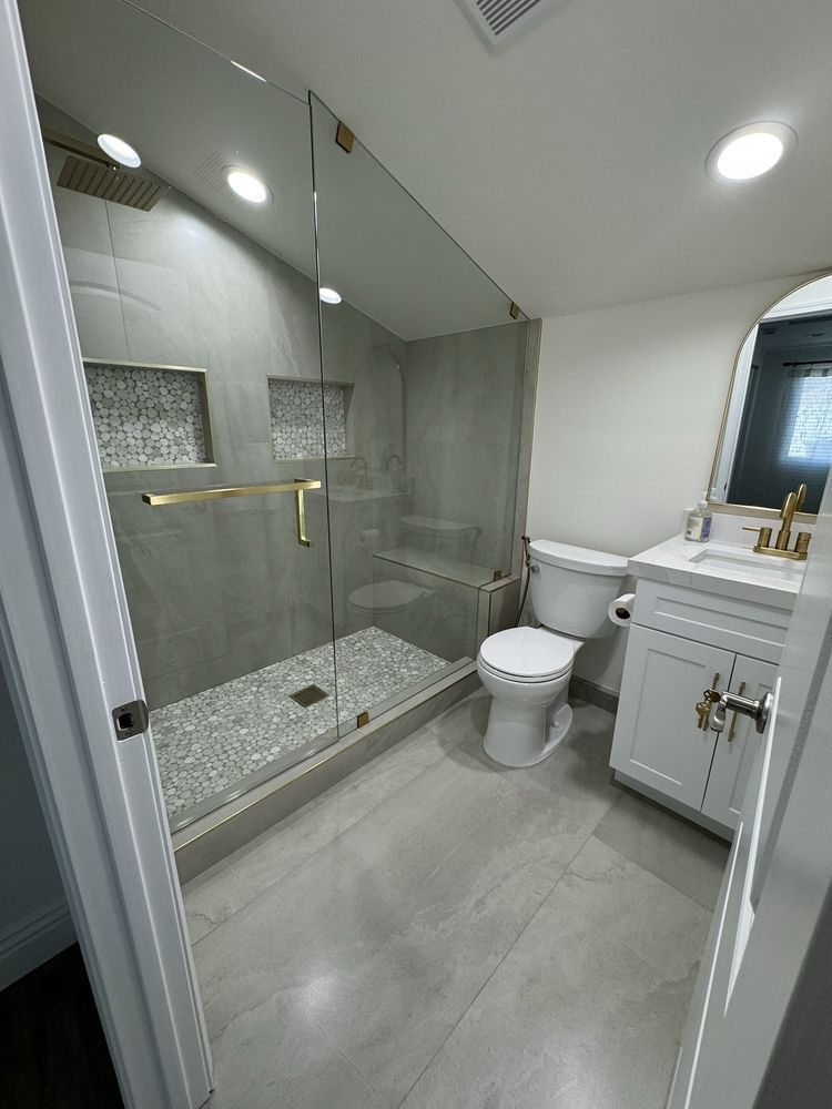 A modern bathroom with a glass-enclosed shower featuring gold hardware, grey tile flooring, and a white vanity.