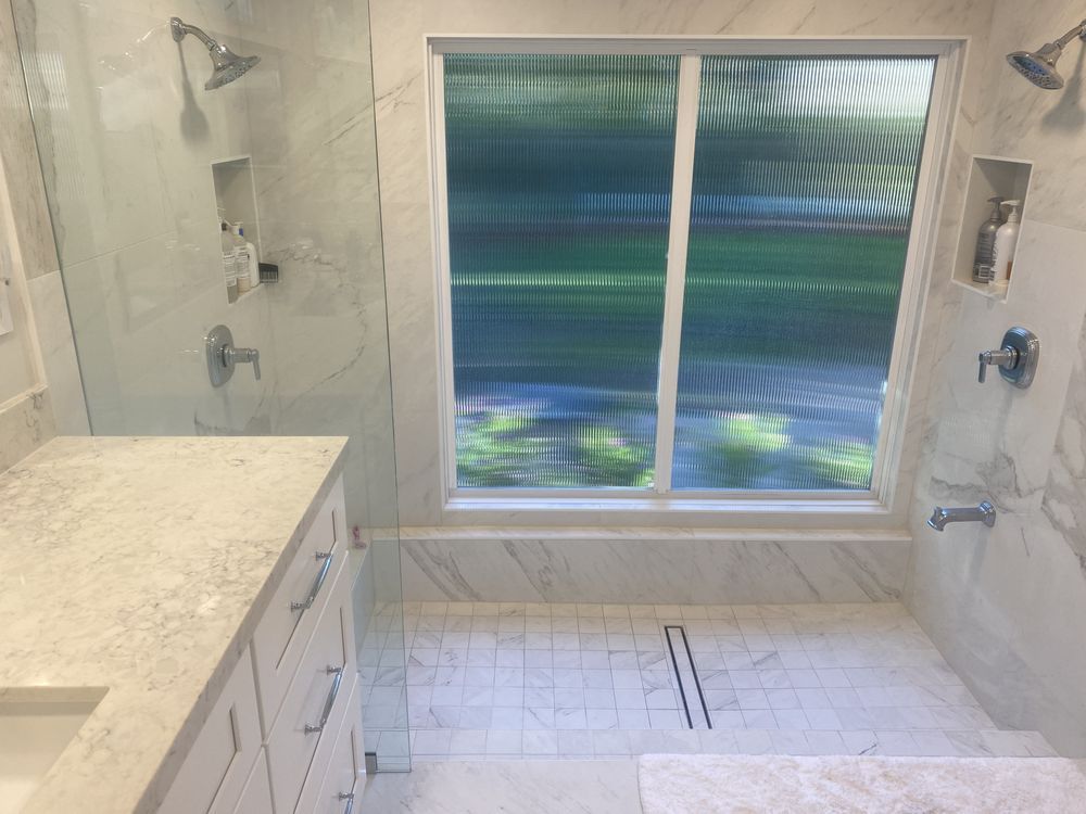 A bright, modern bathroom featuring a walk-in shower with dual showerheads, marble-tiled walls, and a large frosted window.