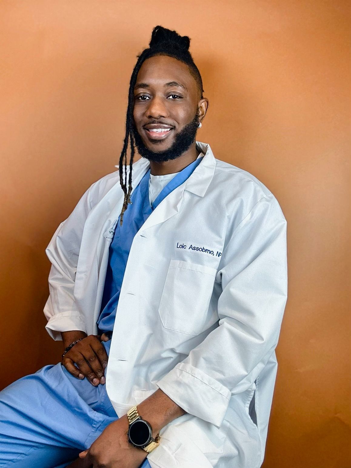 A man wearing a white lab coat and blue scrubs is sitting down.
