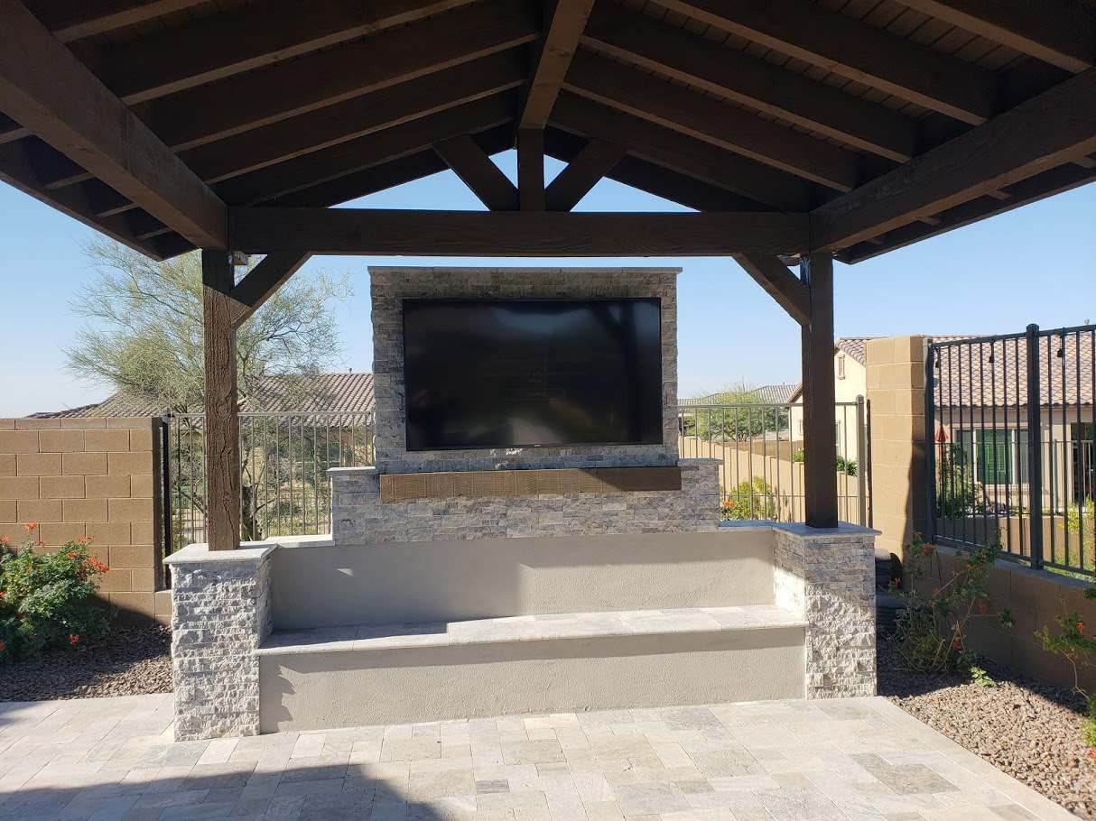 A large flat screen tv is mounted on a stone wall under a wooden gazebo.