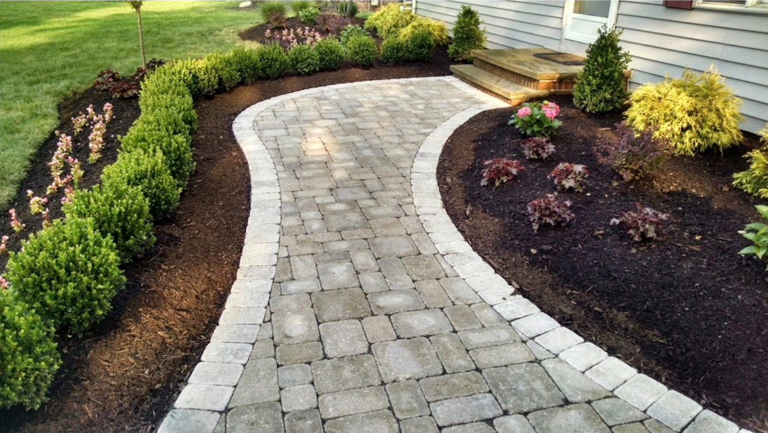 A brick walkway is surrounded by bushes and flowers in front of a house.