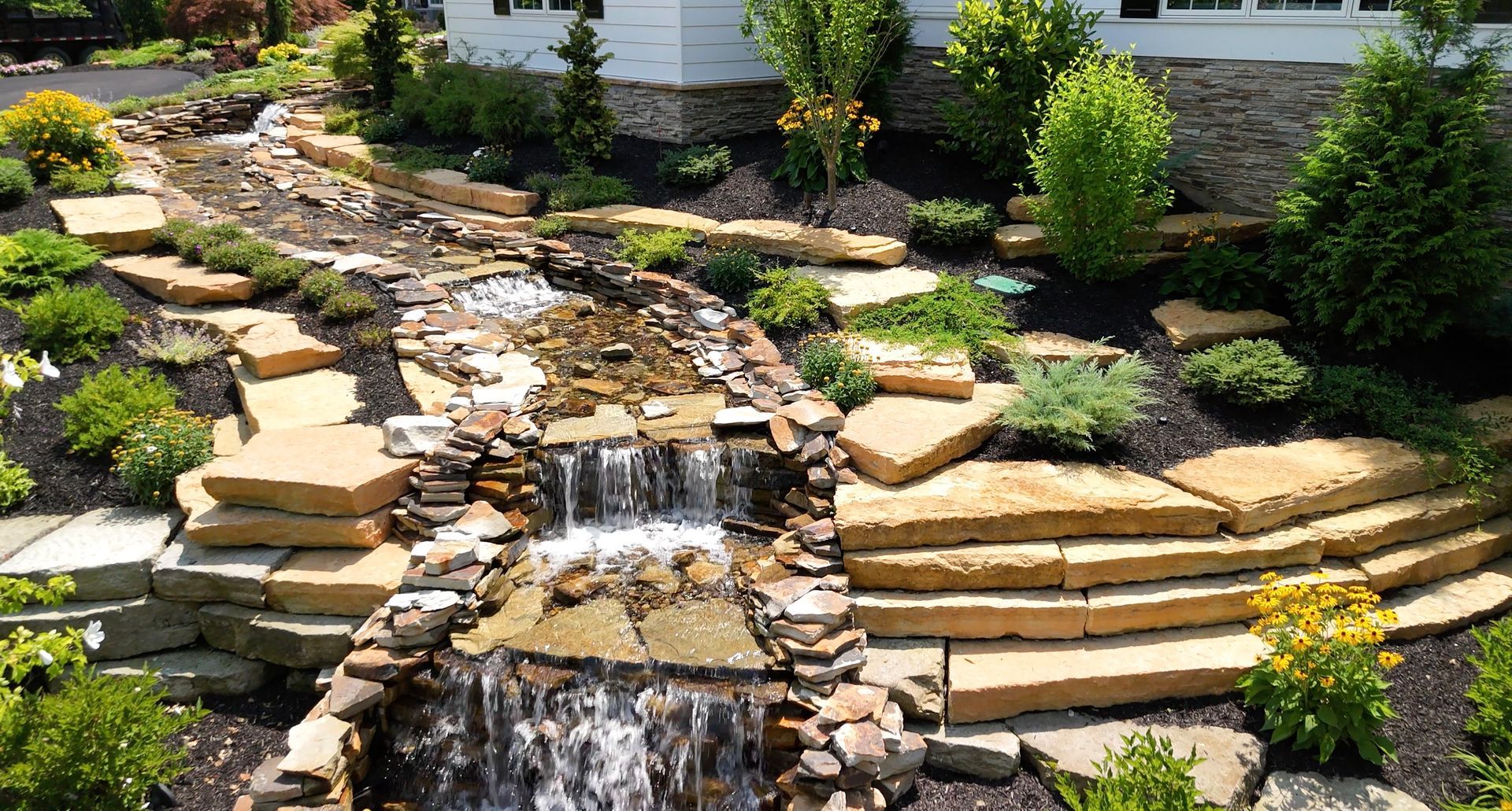 There is a waterfall in the middle of the garden in front of a house.