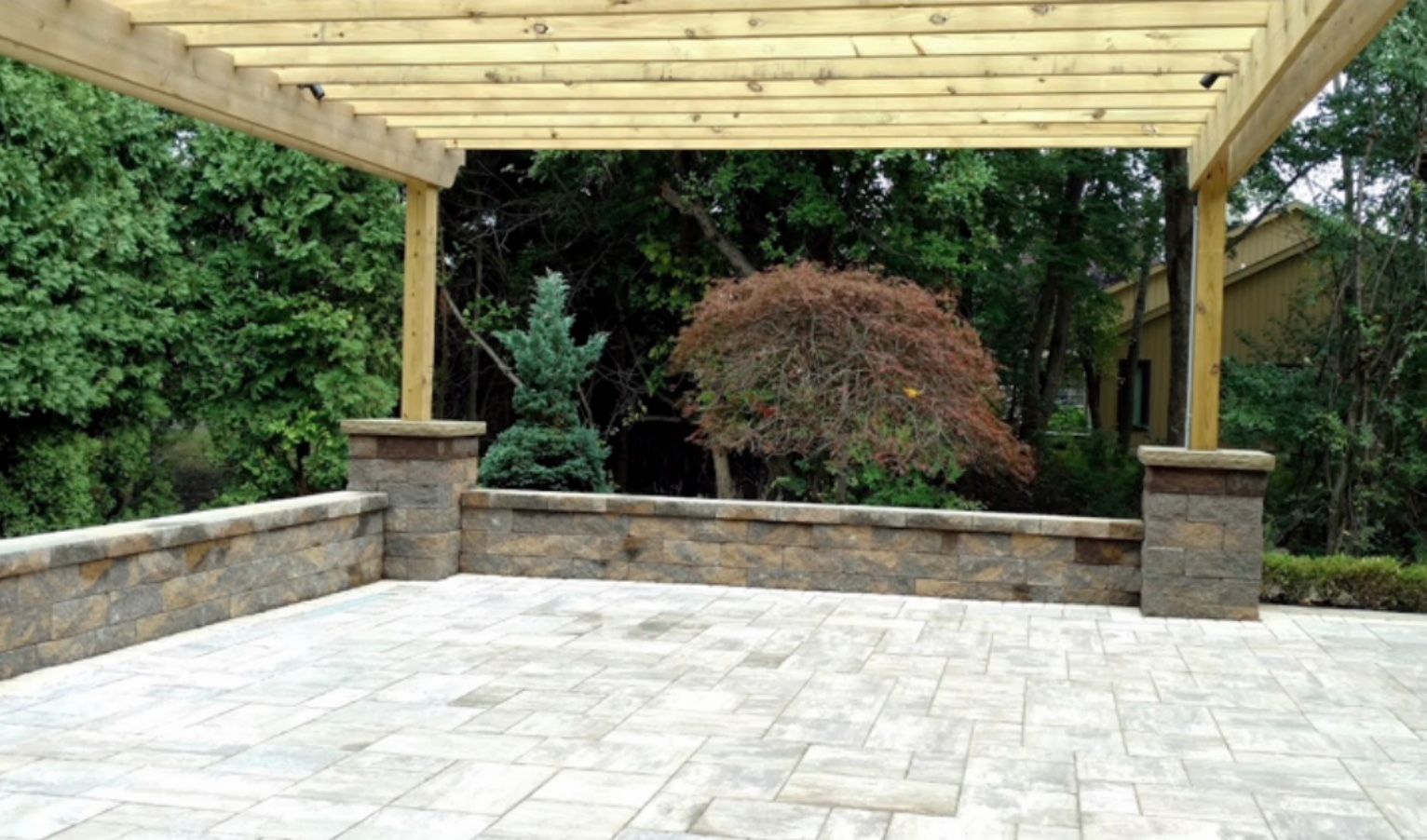 An empty patio with a wooden pergola and trees in the background.