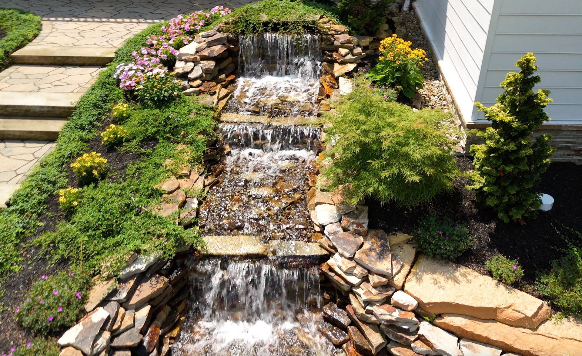 A waterfall is surrounded by rocks and plants in front of a house.
