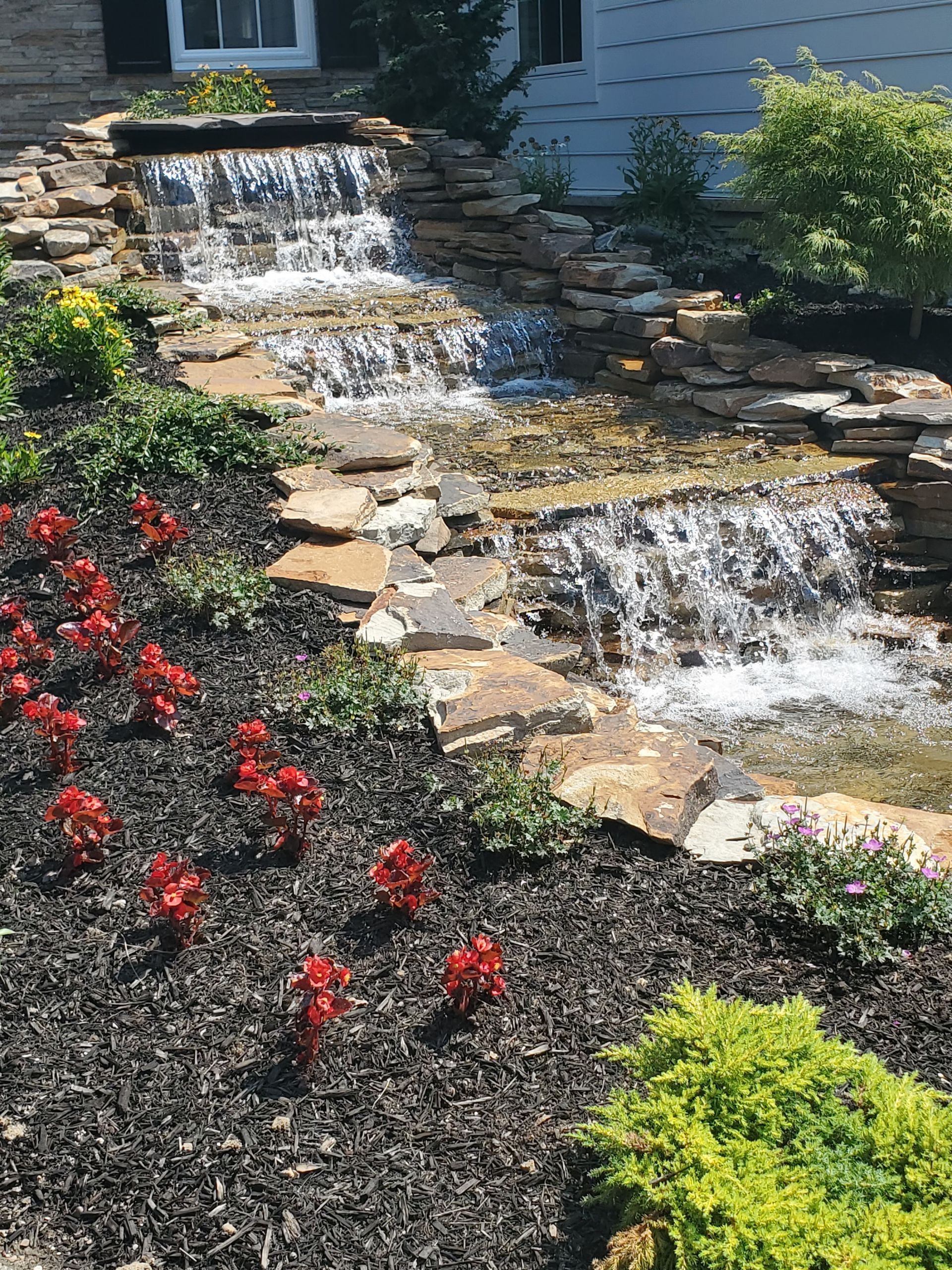 A waterfall is surrounded by flowers and rocks in front of a house.