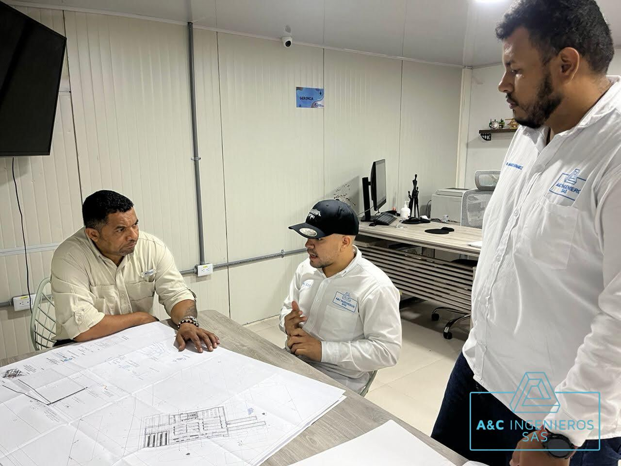 Tres hombres en una habitación revisando planos en una mesa; están vestidos con camisas blancas y tostadas.