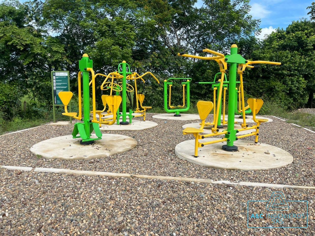 Equipos de fitness al aire libre, de color amarillo y verde, sobre pistas de grava en un parque, con árboles al fondo.