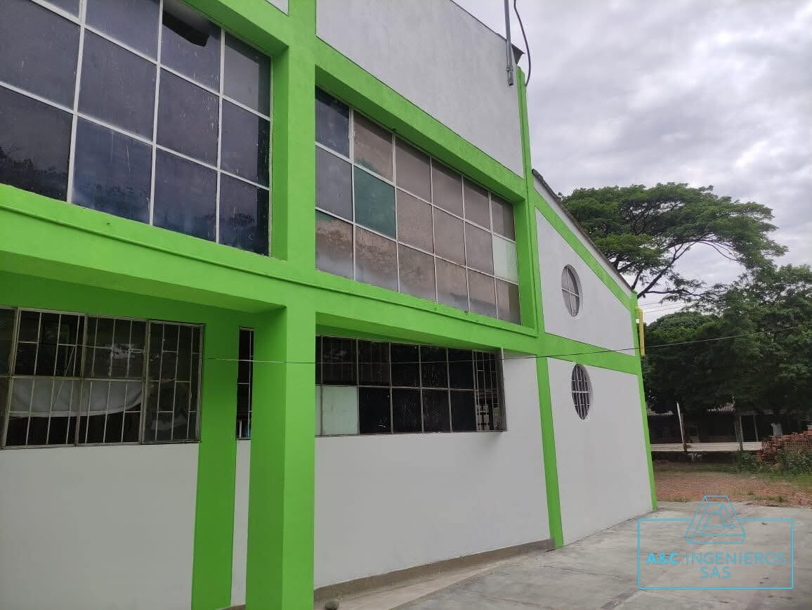 Edificio blanco y verde con grandes ventanales bajo un cielo nublado, cerca de árboles.