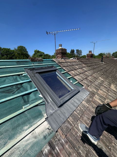 A person on a rooftop installing a skylight on a partially re-tiled roof; clear blue sky overhead.
