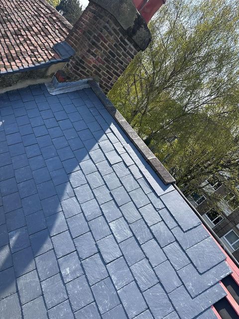 Slate tile roof with a brick chimney and a tree in the background. The tiles are blue-grey.