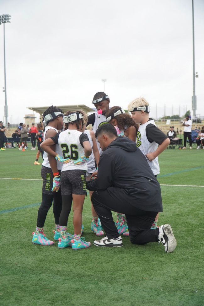 Youth football coach kneeling with players on a turf field during a huddle