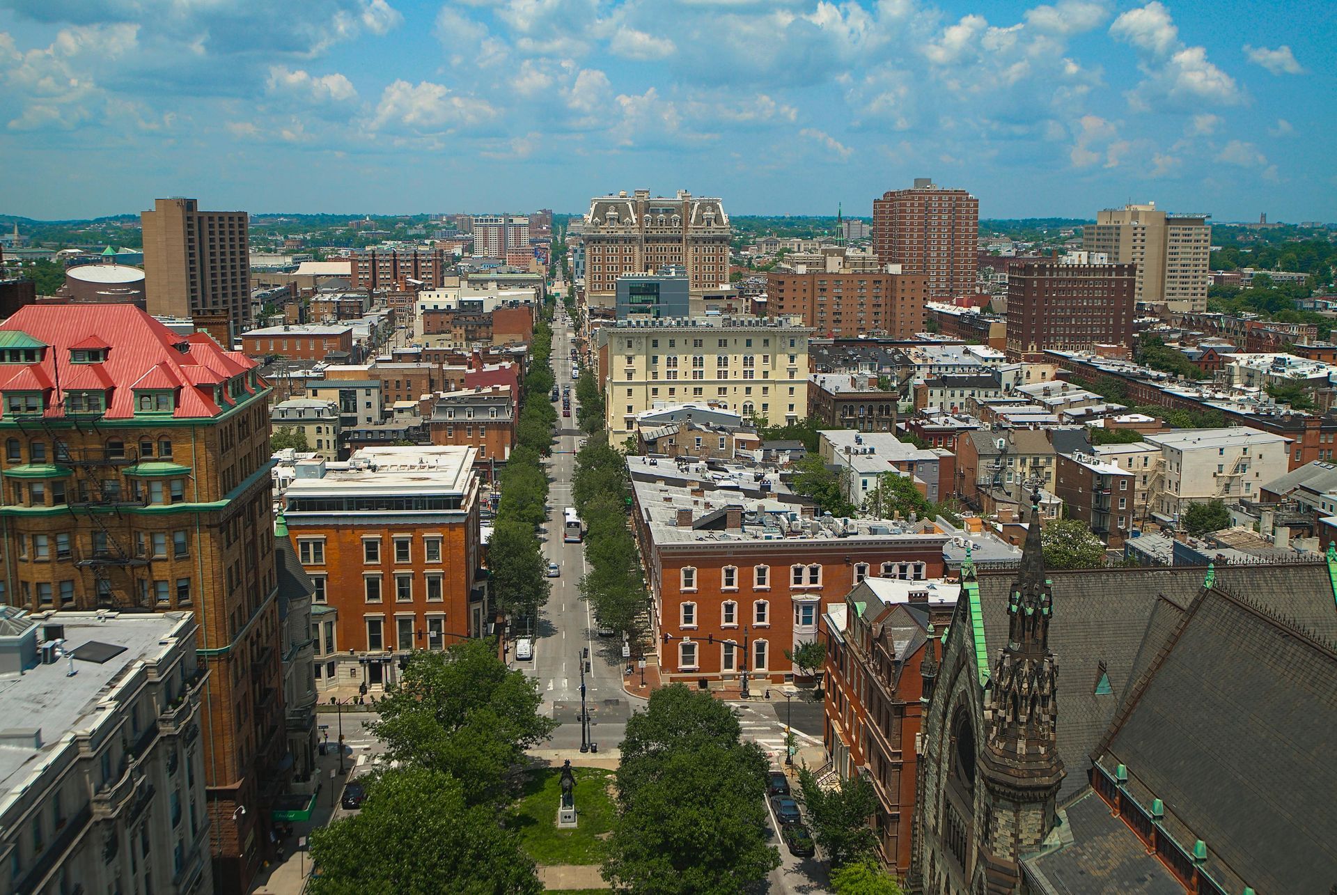 A high-angle view of a city street lined with trees and historic mid-rise brick buildings under a blue, cloudy sky.