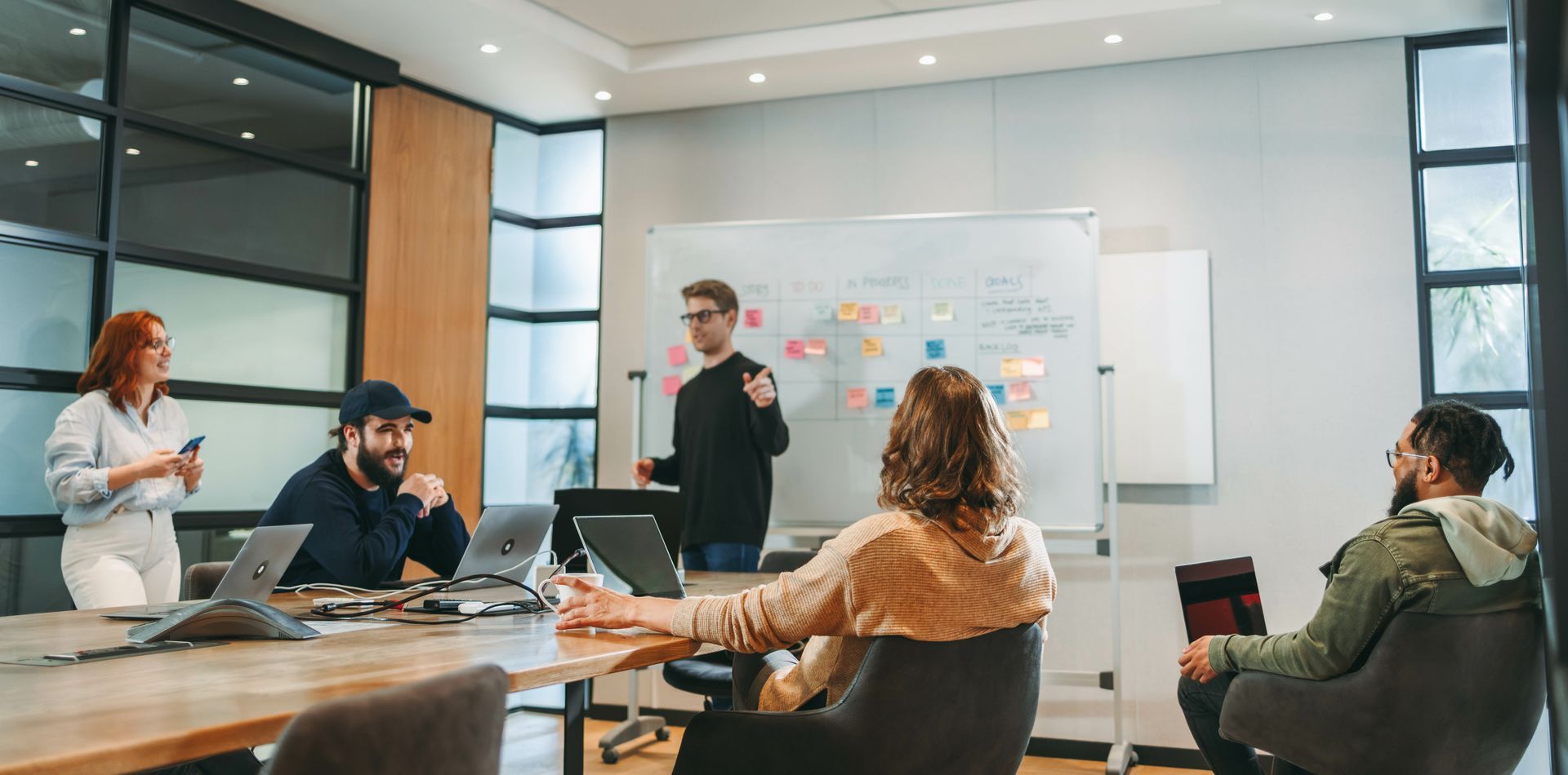 A team meeting in a modern office. A man gestures to a whiteboard as others listen, laptops on the table.