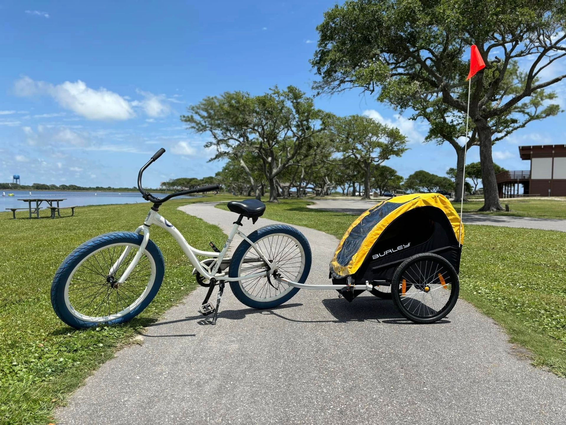 White bicycle with a yellow trailer on a paved path near a body of water under a blue sky.