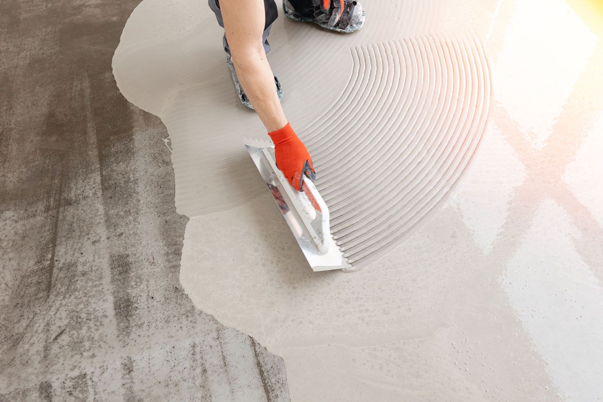 Person using notched trowel to spread adhesive on a concrete floor, preparing for tile installation.
