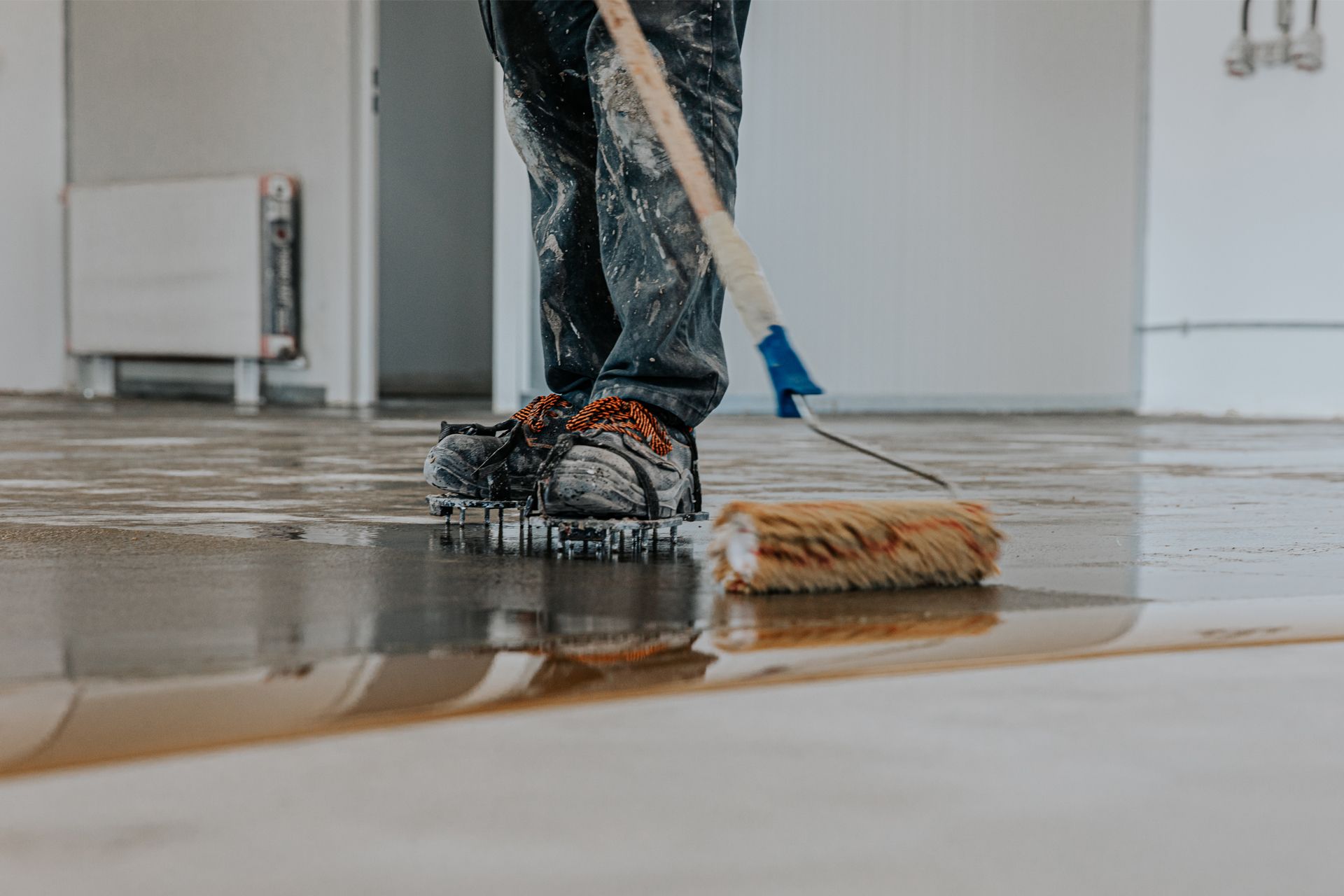 Person applying sealant to floor with roller; splattered paint on work clothes and shoes.
