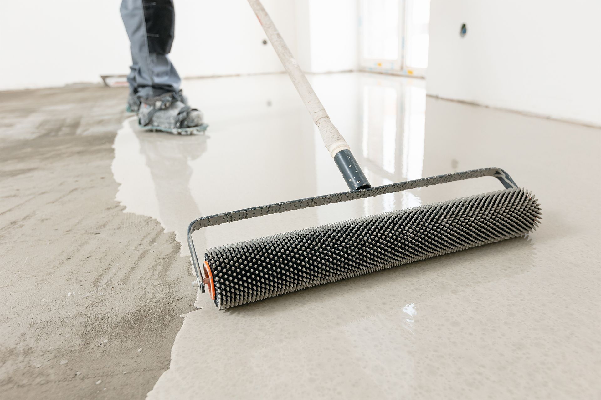 Person rolling a spiked roller over a wet, light gray floor coating.