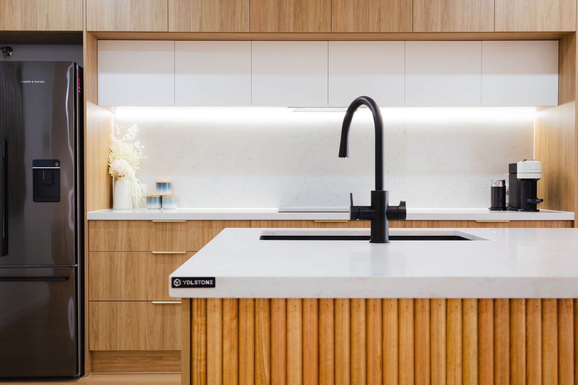 Modern kitchen with a black faucet over a white countertop island with light wood cabinets and a black refrigerator.