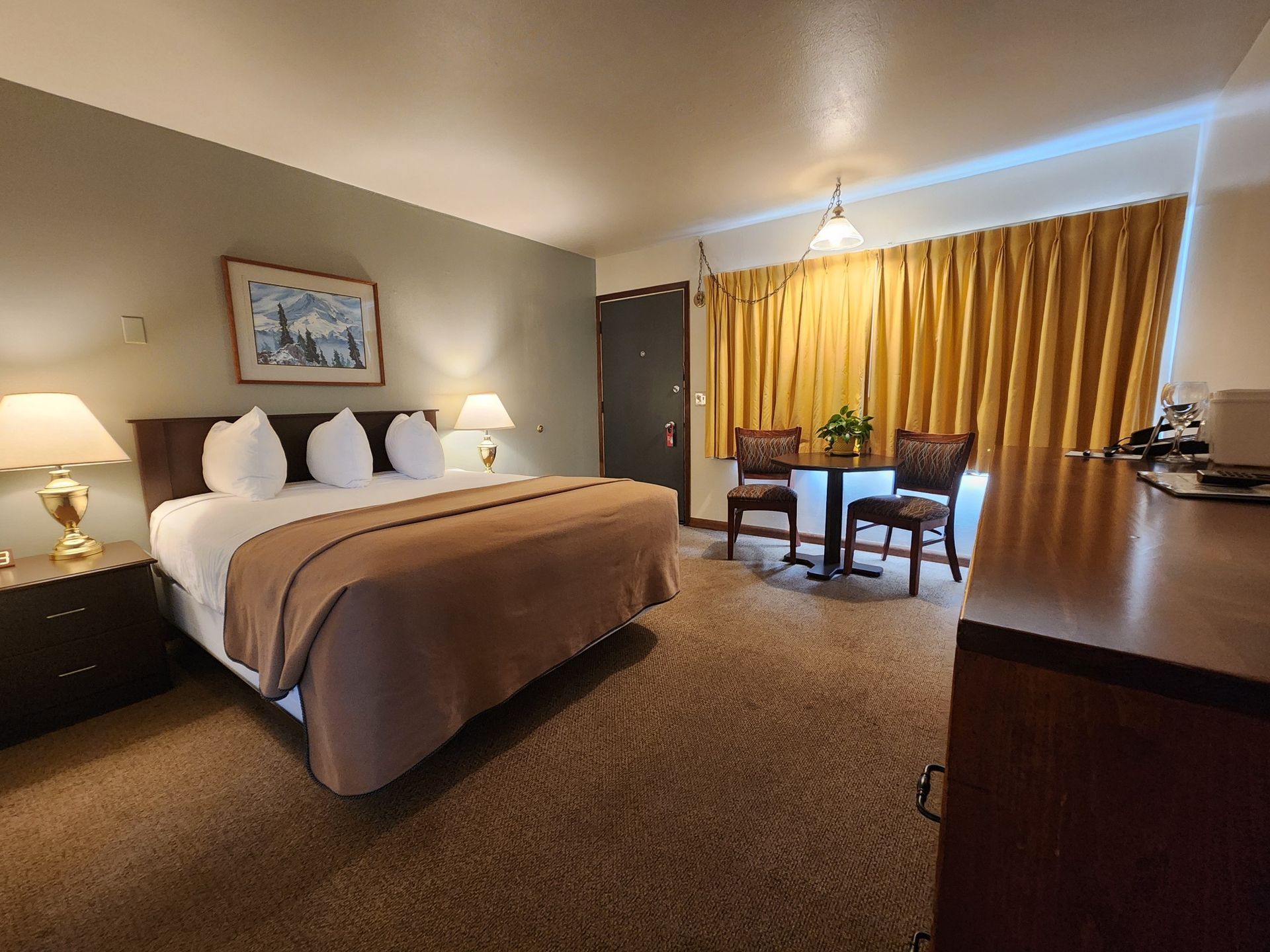 Hotel room interior with a bed, table, and chairs. Brown carpet and curtains.