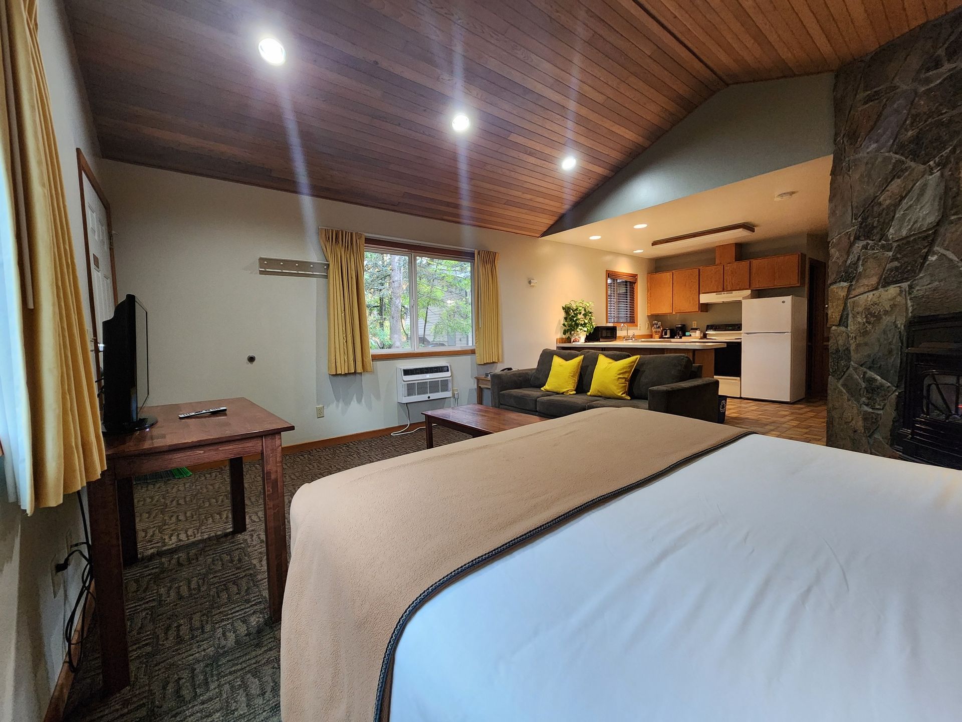 Cozy hotel room with a bed, kitchenette, and fireplace. Warm-toned wood ceiling and stone wall.