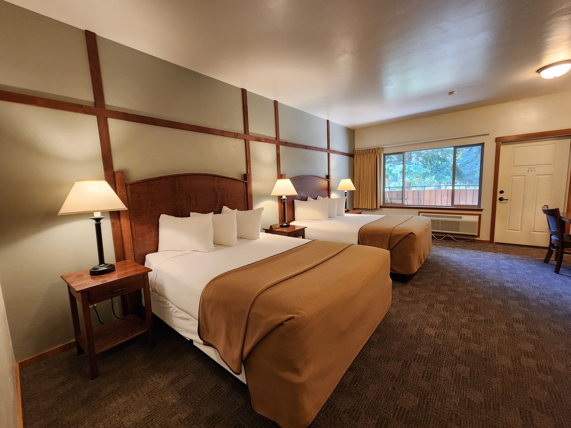 Hotel room with two queen beds, brown blanket, lamps, window, and door.