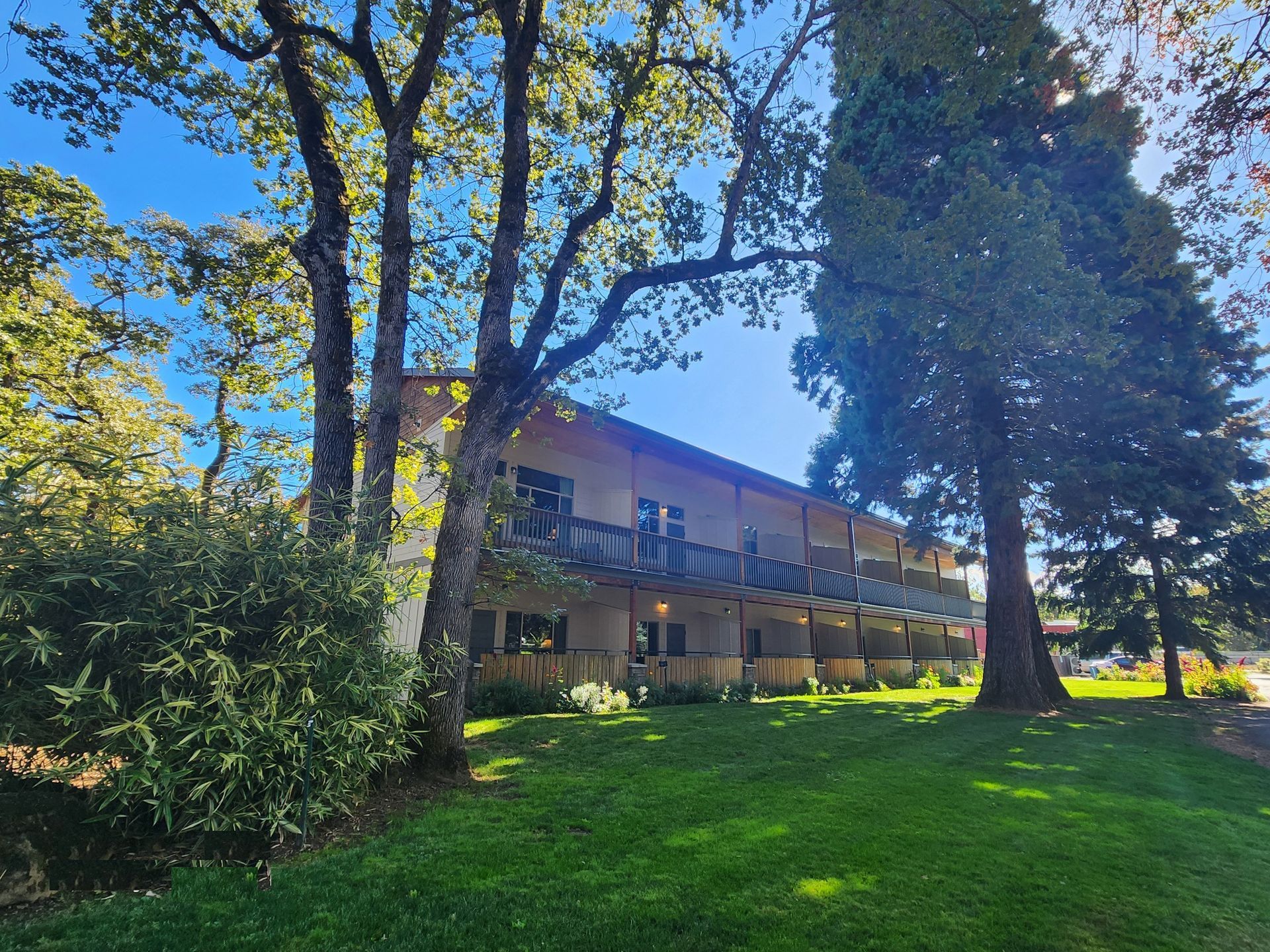 A two-story building with a balcony, trees, and green grass under a blue sky.