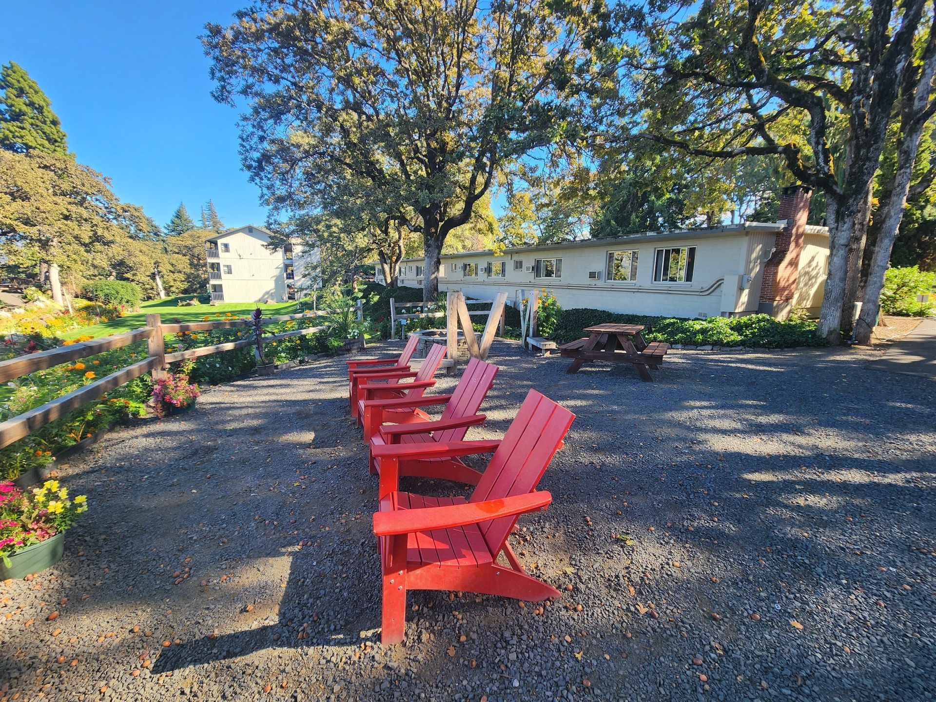 Red Adirondack chairs face a low building with trees and blue sky. Gravel patio with a picnic table.