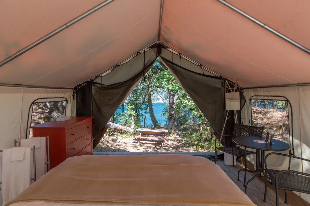 Interior of a glamping tent, with bed, dresser, table, and outdoor view of trees and water.