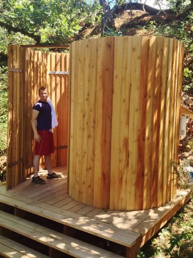 Man stands by a wooden outdoor shower. The shower is cylindrical and open on one side.