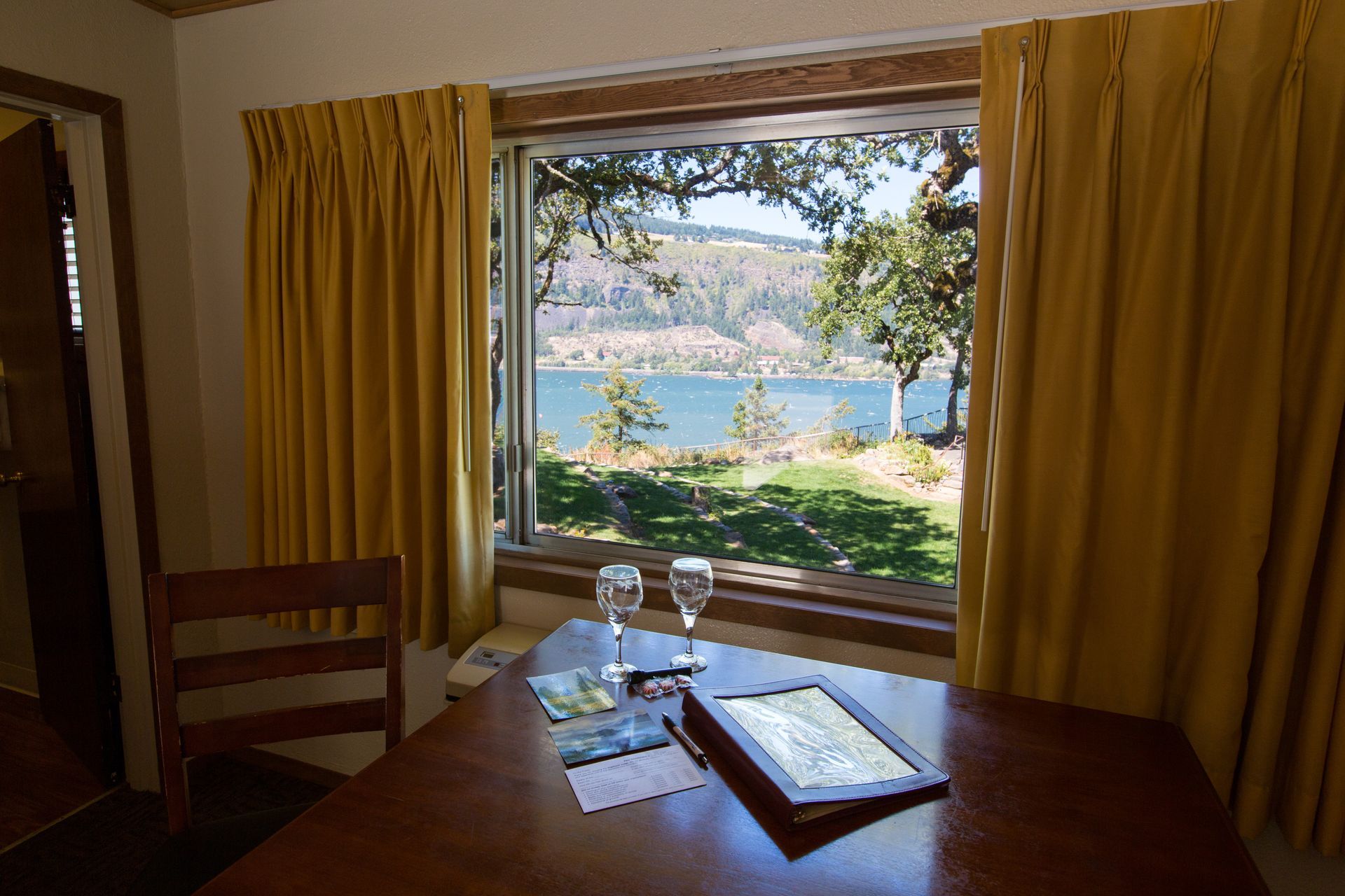 A room with a view: table with champagne flutes, overlooking a lake and trees, framed by yellow curtains.